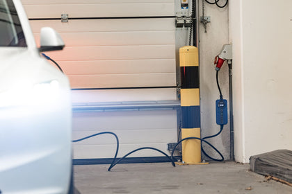 White electric car charging from a wall charger in front of a garage with a blue EV charging cable